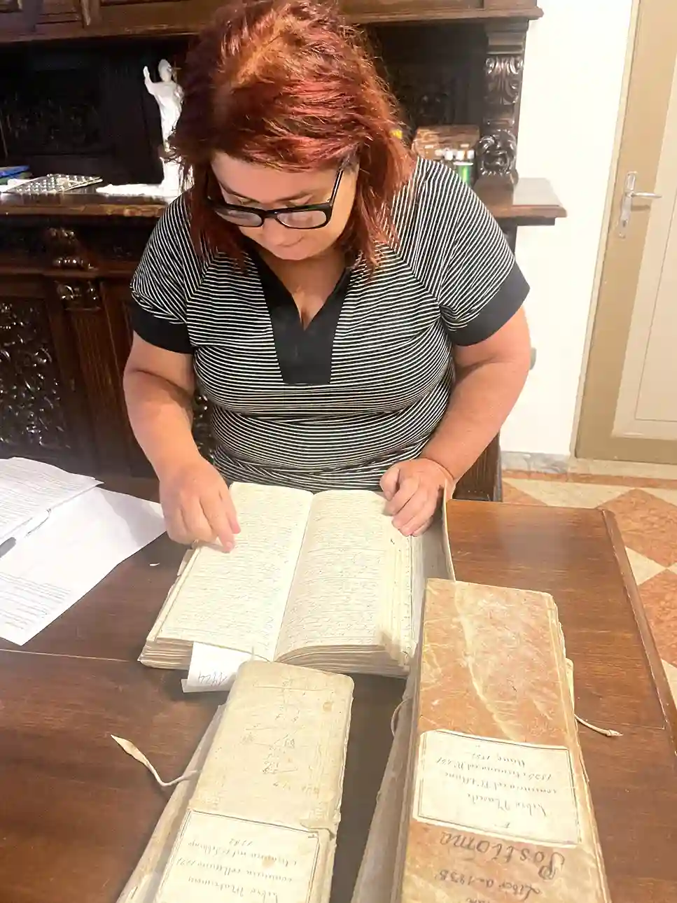Marina Martello standing at a parish table pointing at an open register, with bound volumes labeled Postioma, 1795-1835, on the table
